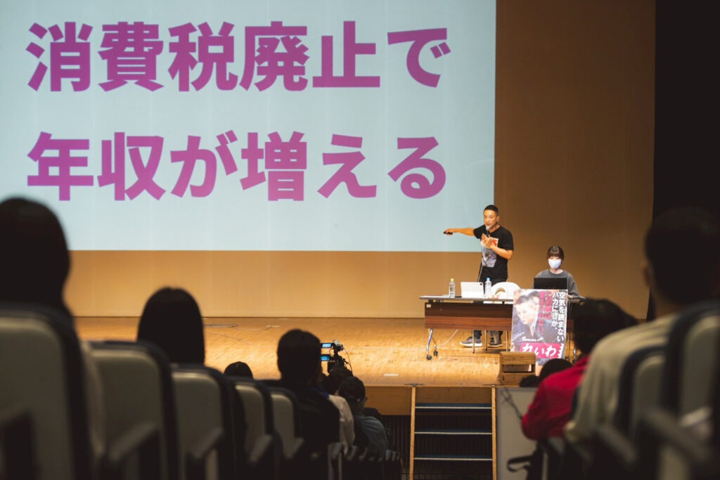 壇上で山本太郎氏が、スクリーン上で「消費税廃止で収入が増える」と訴えている姿